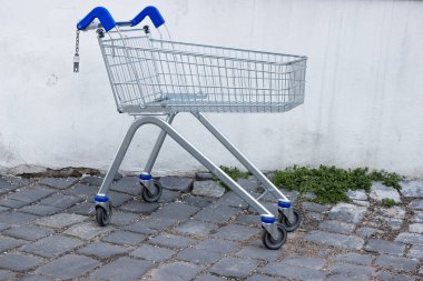 Terk edilmiş, mavi kabzalı boş bir alışveriş arabası kaldırım taşı yolundaki kirli beton duvarın yanında duruyor. Eski kirli grunge duvarları. Kayıp yer. Uzayı kopyala Seçici odak.