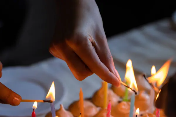 İki kişinin arkadaşı ev yapımı doğum günü pastasında renkli mumlar yakıyor. Kutlama partisi konsepti. Karanlık, bulanık bir arkaplan. Uzayı kopyala Kapatın. Seçici odak.