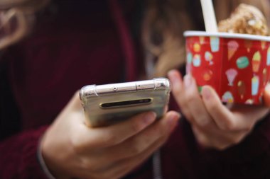 Elinde cep telefonu ve dondurmalı kağıt bardak tutan genç bir kız. Dokunmatik ekran kaydırılıyor. Mesajlaşmak ve dondurma yemek. Seçici odaklanma. Görüntü. Telefona odaklan..