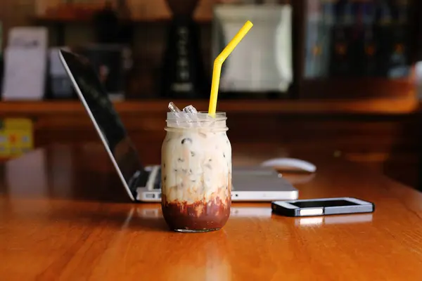 Iced Chocolate separate layers with milk in jar on wooden table have laptop and mobile phone background in cafe. Iced Latte on table with computer notebook and smartphone behind.