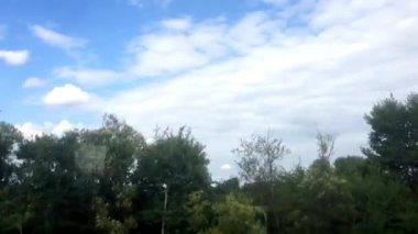 Arka planda birkaç bulut ve ağaç olan açık mavi bir gökyüzü. Gökyüzü çok parlak ve ağaçlar yemyeşil.