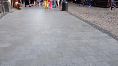 Bir grup insan kaldırımda yürüyorlar. Kaldırım taştan yapılmıştır ve gri bir rengi vardır. Farklı yönlerde yürüyen birkaç kişi var, bazıları el çantası taşıyor.