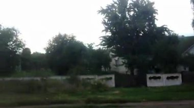 Beyaz çatılı ve mavi kapılı bir ev çitin önünde duruyor. Ev ağaçlar ve çalılarla çevrili.