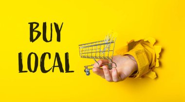 Üzerinde Buy Local yazan bir alışveriş arabasını tutan bir el. Resmin parlak sarı bir arka planı ve sarı ve siyah bir yazı tipi vardır.