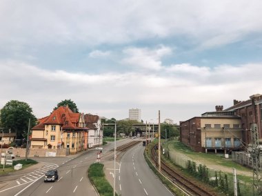 İçinde tren yolu olan bir şehir caddesi. Cadde, bazıları eski ve bazıları daha yeni olan binalarla doludur. Sokaktan geçen arabalar var.