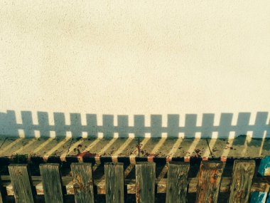 Tahta bir çit beyaz bir duvara gölge düşürür. Çit ahşaptan yapılmıştır ve kırmızı bir şeride sahiptir. Gölge karanlık ve uzamış, duvarın öbür tarafına doğru uzanıyor. Sahne biraz kasvetli.