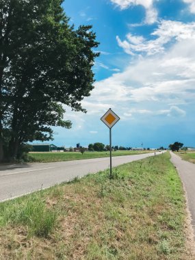 Yol kenarında sarı ve beyaz bir tabela var. Yol boş ve olay yerinde ne araba ne de insan var.