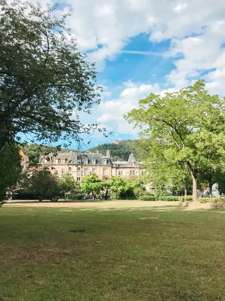 Arkasında büyük bir bina olan bir park. Gökyüzü birkaç bulutla mavidir. Parkta dolaşan insanlar var.