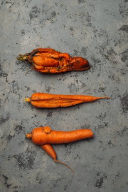 Üç havuç gösteriliyor, biri ikiye bölünmüş ve diğer ikisi bir bütün. Havuçlar gri bir yüzey üzerinde bir sıra halinde düzenlenmiştir. Çirkin sebzeler, yiyecek organik atık azaltma