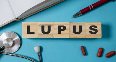 LUPUS harflerinin yazılı olduğu tahta blok. Blok mavi bir yüzey üzerinde, bir kalem ve steteskopla.