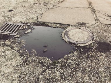 Çatlak bir kaldırımın ortasında bir su birikintisi duruyor. Su birikintisi, yerdeki bir delik tarafından kısmen tıkanmış bir ızgarayla çevrili. Sahne kasvetli ve ıssız.