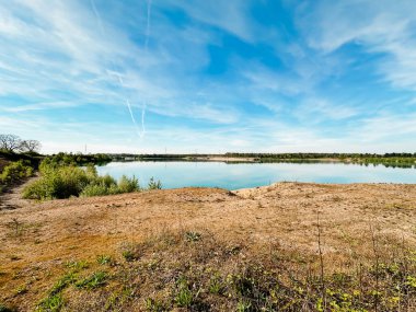 Üzerinde açık mavi gökyüzü olan güzel bir göl. Gökyüzü beyaz bulutlarla kaplı. Göl çimenli bir alanla çevrilidir.