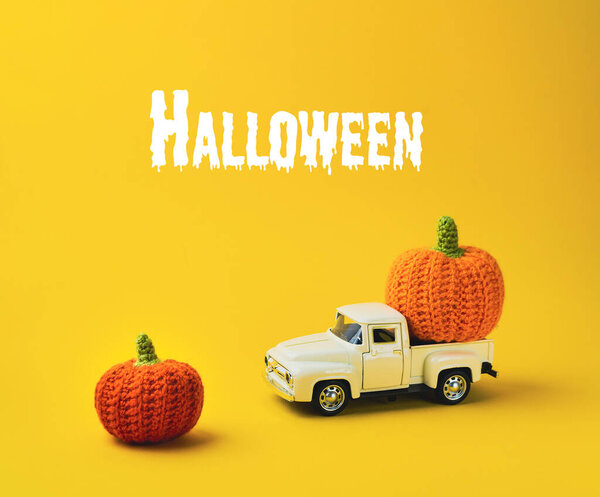 White truck is carrying two orange pumpkins. The pumpkins are placed on the truck bed and the truck is parked on a yellow background