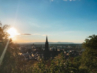 Arka planda büyük bir kilise olan güzel bir şehir manzarası. Güneş batıyor, şehrin üzerine sıcak bir parıltı saçıyor.
