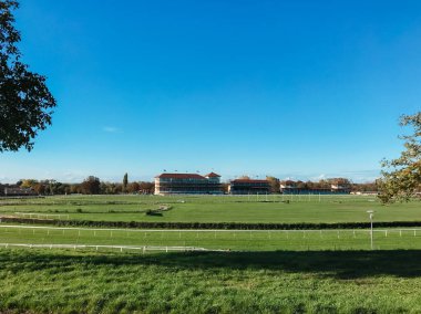 Kırmızı çatılı büyük bir bina bir tarlada yer alır. Gökyüzü açık ve mavi. Binanın etrafı çitlerle çevrili.