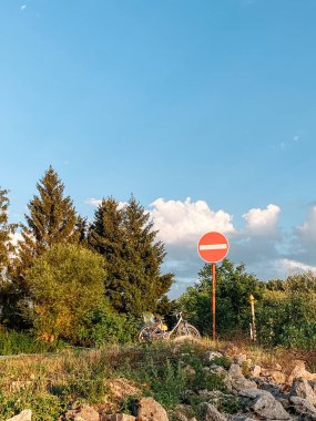 Ortasında beyaz çizgi olan kırmızı ve beyaz tabela bir yamaçta. Tabela ağaçlarla çevrili ve yakınlarda bir bisiklet park edilmiş. Gökyüzü masmavi ve huzur dolu.