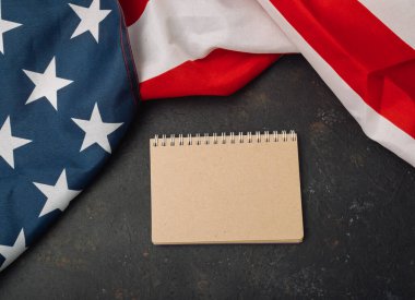 Boş bir defter kırmızı, beyaz ve mavi bir Amerikan bayrağının yanında bir masanın üzerinde duruyor. Bayrak siyah bir yüzeye yayılmış ve defter ortalarına yerleştirilmiş.