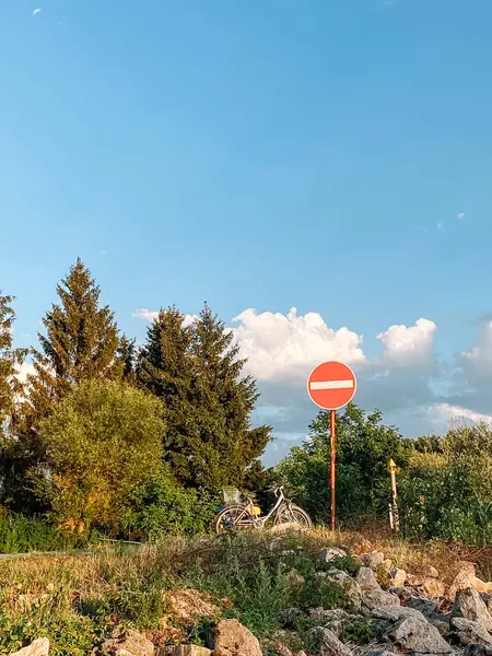 Ortasında beyaz çizgi olan kırmızı ve beyaz tabela bir yamaçta. Tabela ağaçlarla çevrili ve yakınlarda bir bisiklet park edilmiş. Gökyüzü masmavi ve huzur dolu.