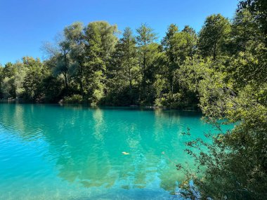 Suyun gövdesi gökyüzüne yansıyor, arka planda ağaçlar var. Su mavi ve sakindir. Huzurlu ve sakin bir atmosfer yaratır.