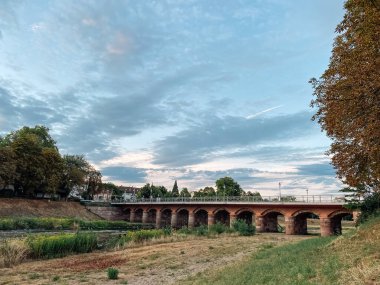 Köprü, arka planda bulutlu bir gökyüzü olan bir nehri gerer. Köprü eski ve kırsal bir görünüme sahip.