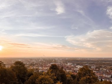 Gökyüzünde birkaç bulut olan bir şehrin üzerinde güzel bir gün batımı. Gökyüzü mavi ve turuncunun bir karışımıdır, sıcak ve huzurlu bir atmosfer yaratır. Aşağıdaki şehir hareketlilikle dolup taşıyor
