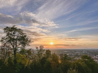 Gökyüzü birkaç bulutla mavidir ve güneş batıyor. Ağaçlar yeşil ve manzarası bir şehir.