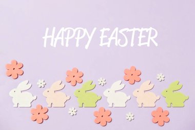 Mor arkaplanlı renkli Paskalya tavşanı süslemesi. Tavşan süslemeleri sıraya dizildi ve arka planda çiçekler var. Mesaj şu: Mutlu Paskalyalar.