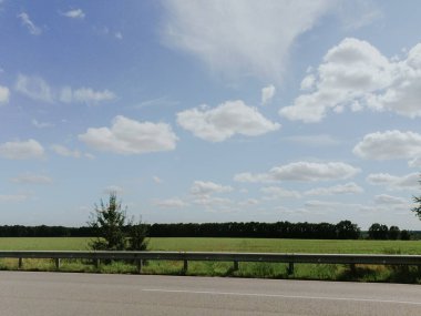 Yan tarafında çit, ortasında ağaç olan uzun bir yol. Gökyüzü açık ve mavidir birkaç bulutla