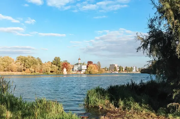 Arka planda yeşil ağaç olan bir su kütlesi. Su sakin ve berrak. Uzakta bazı binalar var.