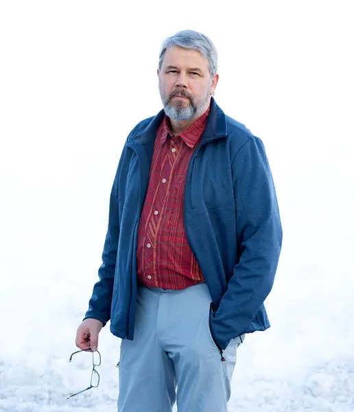 Man, 56 years old with gray hair and a gray beard, standing in front of a snowy area in winter, wearing a red checked shirt, dark blue jacket, light gray trousers, glasses in his hand, one hand in his trouser pocket