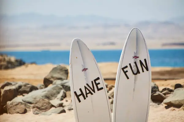 two used surfboards used as a sign saying Have Fun on a beach. sand, rocks and ocean in the background