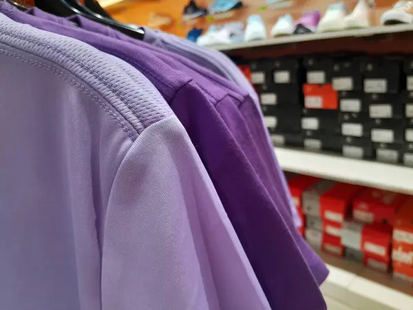 Arka planda ayakkabı rafları olan bir mağazada askıdaki mor spor tişörtleri..