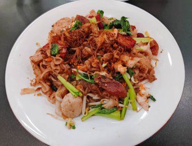 Çin yemeği Kızarmış Düz Pirinç Eriştesi (kway teow), karides, sığır köftesi, lapa cheong veya Çin sosisi ve siyah arka planda izole edilmiş beyaz tabak üzerine kırmızı domuz eti..