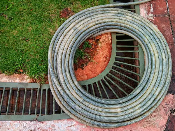 Yuvarlak kanalizasyon kapısına, çimenlere ve kaldırıma dayanan sarmalanmış bahçe hortumunun üst görüntüsü..