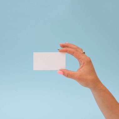 Renkli manikürlü, mavi stüdyo duvarında izole edilmiş boş bir kağıt kart. Doğal ışıklandırma ve aralıklar net görsel mesaj ve ticari adaptasyon oluşturur.