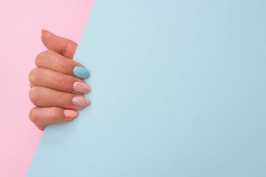 Kağıt çarşaf tutan parlak pembe ve mavi tırnaklı şık bir el. Renkli arka planda pastel renklerle çekiliyor. Manikür ve simetri göz alıcı denge yaratır.
