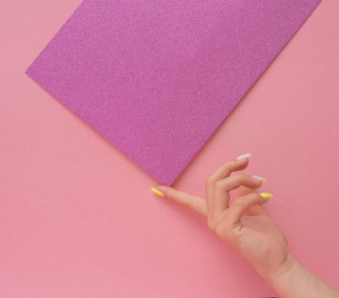Pastel tırnakları olan narin bir kadın eli pembe arka planda parlak mor bir örtüyü dengeliyor. Tasarımı, zarafeti ve sanatsal ahengi vurgulayan minimalist düz uzanma.
