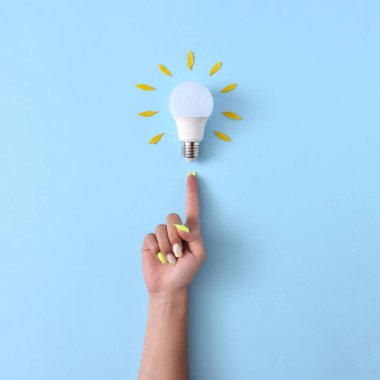Sarı yapraklı, parlak fikirleri ve yeniliği sembolize eden yaratıcı bir ampul konsepti. Pastel mavi arka plandaki ampulü gösteren sarı tırnaklı kadın eli..