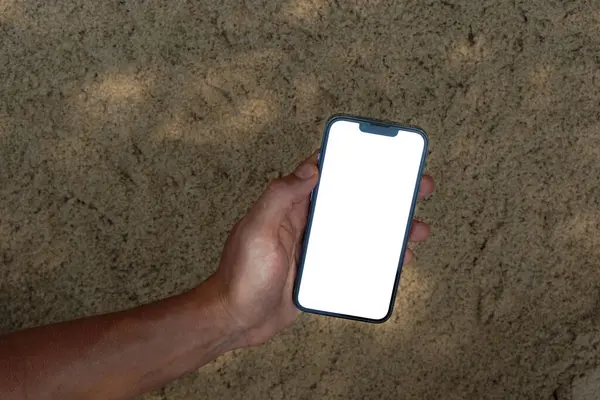 Yazın kumsaldaki kumların üzerinde beyaz, boş bir telefon ekranı tutuyor.