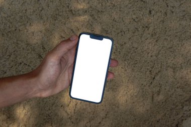 Yazın kumsaldaki kumların üzerinde beyaz, boş bir telefon ekranı tutuyor.