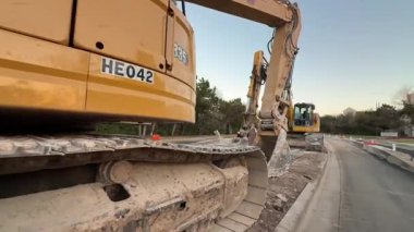 Video, ağır makineli bir inşaat sahnesini gösteriyor, toprak bir yolda sarı bir kazıcı ile başlıyor, sonra buldozerler ve daha fazla kazıcının kazdığı