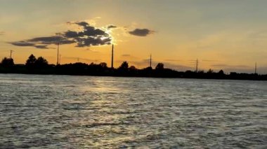 Suyun üzerinde nefes kesen bir günbatımı canlı turuncudan, güneş batarken derin çivit mavisi rengine dönüşür. Şehrin silueti, sahnenin karşısında yer alır.