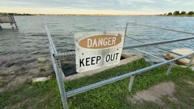 Huzurlu bir su kütlesinin yanındaki tehlike işareti yoldan geçenleri uzak durmaları konusunda uyarıyor, insan uyarıları ile doğal güzellikler arasındaki farkı vurguluyor.
