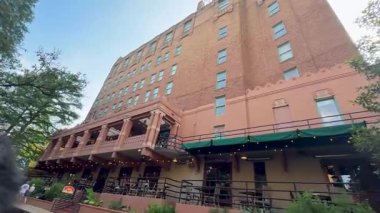 Bu video yeşil tenteli, masalı, sandalyeli ve nehre bakan balkonlu büyük bir tuğla bina içerir.