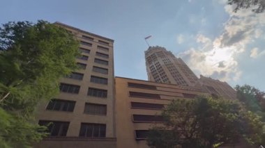17 Mayıs 2025, San Antonio Teksas, ABD - Video, ağaçlarla çerçevelenmiş, bayrağı olan yüksek bir binayı gösteriyor