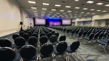 Kalabalık bir konferans salonu, seyirci sıra sıra sandalyeleri doldururken, etkinliğin önemini vurgulayarak bomboştu.