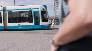 Amsterdam, Hollanda 29 Temmuz 2025: Canlı bir şehir caddesinde yürüyen tesettürlü bir kadın mavi ve beyaz bir tramvay süzülerek şehir yaşamını vurguluyor.