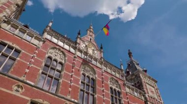 Video klasik saat kulesi olan büyük bir kırmızı tuğla binayı gösteriyor. Önde bir gökkuşağı bayrağı dalgaları, çeşitliliği ve dahil edilmeyi simgeliyor.