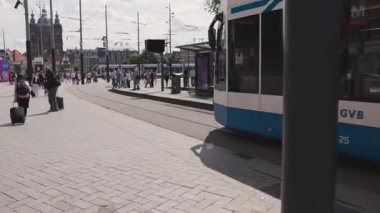 Amsterdam, Hollanda 29 Temmuz 2025: Arka planda çağdaş ulaşım altyapısı ve tarihi mimarinin yer aldığı, yolcu ve bavullarla dolu canlı bir şehir tramvay istasyonu.