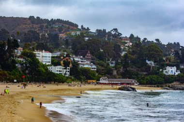 Zapallar sahilinde çok az insan var. Yakınlarda çok sayıda ev ve ağaç var. Bulutlu bir gün.